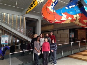 A group of smiling Bagels & Bytes attendees standing under the colorful oversized Crane (water bird) in the Children's Museum lobby.